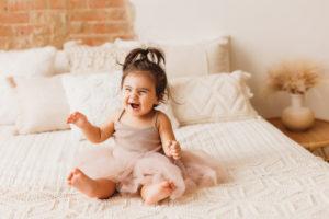 milestone photographer, a toddler girl is happy in her dress, sitting on the bed