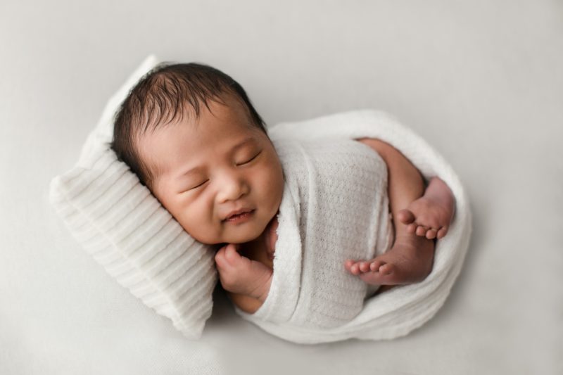 newborn boy smiling swaddled in white laying on pillow